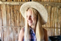 Female teenager in straw hat smiling and looking away while standing against wooden partition inside shed on farm — Stock Photo