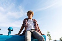 Хлопчик в повсякденному вбранні дивиться на камеру, сидячи на синьому тракторі проти хмарного неба в сонячний день на фермі — стокове фото