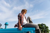 Seitenansicht eines Jungen im lässigen Outfit, der in die Kamera blickt, während er an einem sonnigen Tag auf einem Bauernhof auf einem blauen Traktor vor bewölktem Himmel sitzt — Stockfoto
