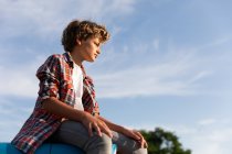 Seitenansicht eines Jungen in lässigem Outfit, der an einem sonnigen Tag auf einem Bauernhof auf einem blauen Traktor vor bewölktem Himmel sitzt und wegschaut — Stockfoto