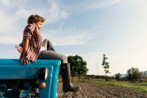 Vista lateral del niño en traje casual mirando hacia otro lado mientras está sentado en el tractor azul contra el cielo nublado en el día soleado en la granja - foto de stock