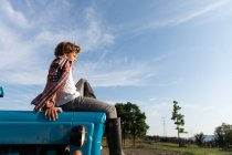 Вид сбоку мальчика в повседневной одежде, который смотрит в сторону, сидя на голубом трамвае на фоне облачного неба в солнечный день на ферме — стоковое фото