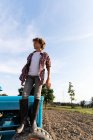 Side view of boy in casual outfit looking away while standing on blue tractor against cloudy sky on sunny day on farm — Stock Photo