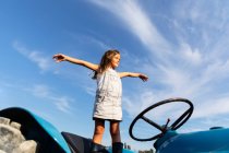 Little girl in denim dress stretching out arms while standing on tractor against cloudy sky on farm — Stock Photo