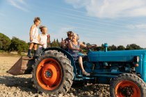 Ältere Frau fährt Traktor und sieht Kinder bei der Arbeit auf dem Acker am sonnigen Tag auf dem Bauernhof — Stockfoto