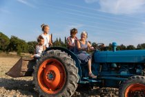 Ältere Frau fährt Traktor und sieht Kinder bei der Arbeit auf dem Acker am sonnigen Tag auf dem Bauernhof — Stockfoto