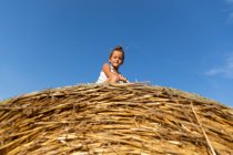 Kleines Mädchen blickt in die Kamera, während es an einem sonnigen Tag auf einem Bauernhof auf trockenem Gras vor wolkenlosem blauem Himmel sitzt — Stockfoto