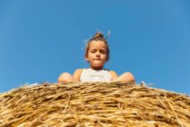Маленькая девочка смотрит в камеру, сидя на сухой траве против безоблачного голубого неба в солнечный день на ферме — стоковое фото