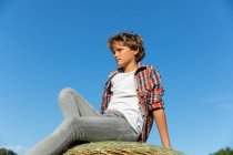 Young boy looking at away while sitting on roll of dry grass against cloudless blue sky on sunny day on farm — Stock Photo