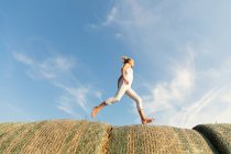 Босоногая девушка, бегущая по рулонам сухой травы на фоне облачного голубого неба в солнечный день на ферме — стоковое фото