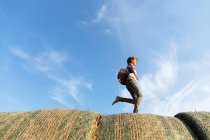 Вид збоку босоніж хлопчик біжить на рулонах сушеної трави проти похмурого блакитного неба в сонячний день на фермі — стокове фото