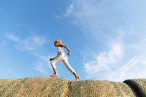 Vista laterale della ragazza scalza che corre su rotoli di erba secca contro il cielo blu nuvoloso nella giornata di sole in fattoria — Foto stock