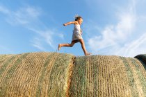 Seitenansicht des barfüßigen Mädchens läuft auf Rollen aus getrocknetem Gras gegen bewölkten blauen Himmel an einem sonnigen Tag auf dem Bauernhof — Stockfoto