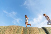 Side view of three kids running on rolls of straw together against cloudy blue sky in agricultural field — Stock Photo