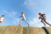 Vista lateral de tres niños corriendo en rollos de paja juntos contra el cielo azul nublado en el campo agrícola - foto de stock