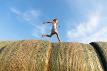 Вид збоку босоніж дівчина біжить на рулонах сушеної трави проти похмурого блакитного неба в сонячний день на фермі — стокове фото