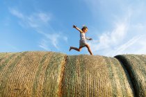 Side view of barefoot girl running on rolls of dried grass against cloudy blue sky on sunny day on farm — Stock Photo