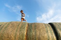 Side view of barefoot girl running on rolls of dried grass against cloudy blue sky on sunny day on farm — Stock Photo