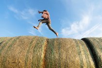 Вид сбоку босоногого мальчика, бегущего по рулонам сухой травы против облачно-голубого неба в солнечный день на ферме — стоковое фото