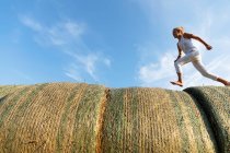 Vista lateral de la chica descalza corriendo sobre rollos de hierba seca contra el cielo azul nublado en el día soleado en la granja - foto de stock