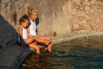 Vista lateral de duas meninas sentadas perto de parede rasgada e salpicando água limpa de lagoa artificial no dia ensolarado — Fotografia de Stock