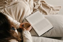 Неузнаваемая женщина сидит на кровати, читает книгу и пьет кофе. — стоковое фото