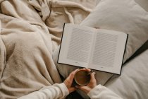 Unerkennbare Frau sitzt auf dem Bett, liest ein Buch und trinkt Kaffee — Stockfoto
