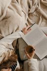 Donna irriconoscibile seduta sul letto a leggere un libro e bere caffè — Foto stock