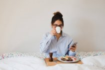 Портрет веселої жінки, що сидить на ліжку з чашкою в руках і підносять здорову їжу на ногах при використанні смартфона — стокове фото