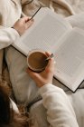 Неузнаваемая женщина сидит на кровати, читает книгу и пьет кофе. — стоковое фото