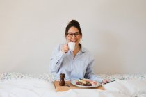 Портрет веселой женщины, сидящей на кровати с чашкой в руках и подносом со здоровой пищей на ногах при использовании смартфона — стоковое фото