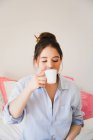 Verträumt schöne junge Frau trinkt erfrischenden Kaffee aus Becher, während sie mit geschlossenen Augen auf dem Bett sitzt — Stockfoto