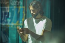 Молодий бородатий красивий чоловік спирається на стіну з мобільним телефоном в руках вдумливо смс — стокове фото