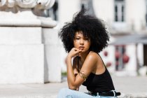 Ethnic woman in jeans and tank top relaxing and sunbathing on stone stairs against urban background — Stock Photo