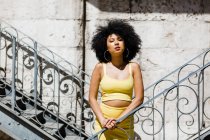 Mujer afroamericana en traje amarillo de pie y apoyada sobre barandilla sobre fondo urbano - foto de stock