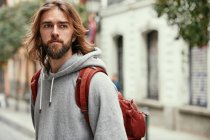 Homem bonito barbudo jovem com capuz cinza com mochila andando seriamente fora — Fotografia de Stock