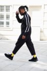 Vue latérale de l'homme afro-américain avec des cornrows en costume de sport noir marchant sur fond urbain — Photo de stock