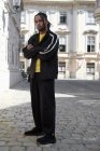 Jeune homme ethnique aux cheveux tressés portant un costume de sport noir regardant la caméra sur fond urbain — Photo de stock
