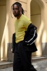 Jovem homem étnico com cabelo trançado vestindo terno esportivo preto olhando para a câmera no fundo urbano — Fotografia de Stock