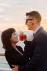 Vista lateral de jovem casal atraente beber bebida vermelha com palhas de um copo no fundo do mar por do sol — Fotografia de Stock