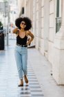 Joven mujer étnica en jeans y camiseta sin mangas caminando y sonriendo a la cámara al aire libre - foto de stock