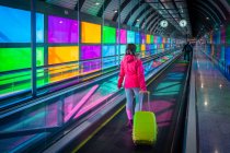 Невпізнанний жінка з валізою їзда косіння доріжки біля барвистих панелей усередині аеропорту Мадрид Барахас в Іспанії — стокове фото