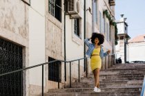 Mujer afroamericana en traje amarillo y chaqueta de mezclilla caminando abajo y mirando a la cámara en el fondo urbano - foto de stock