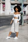 Donna afroamericana sorridente in abito bianco alla moda in piedi con mano in tasca sul ciglio della strada sullo sfondo urbano — Foto stock