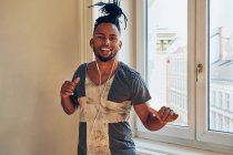 Homme afro-américain souriant avec des tresses dansant sur la musique avec des écouteurs à la maison sur fond de fenêtre — Photo de stock