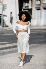 Mulher afro-americana em branco roupa de moda em pé com as mãos em bolsos na beira da estrada contra o fundo urbano — Fotografia de Stock