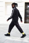 Vue latérale de l'homme afro-américain avec des cornrows en costume de sport noir marchant sur fond urbain — Photo de stock