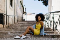 African American woman in yellow suit and denim jacket looking at camera while sitting on stairs on urban background — Stock Photo