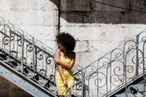 Mulher afro-americana de terno amarelo em pé perto de trilhos e olhando para o fundo urbano — Fotografia de Stock
