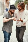 Hombre guapo en sombrero negro de pie con un amigo en la calle y apuntando al teléfono móvil - foto de stock